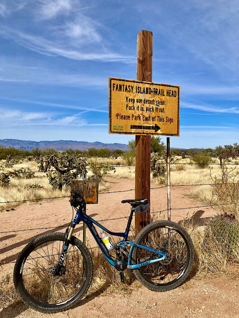 Blue mountain bike leaning against barbed wire fence and post with sign reading: Fantasy Island Trailhead. Keep our desert clean. Pack it in, pack it out. Please part east of this sign.