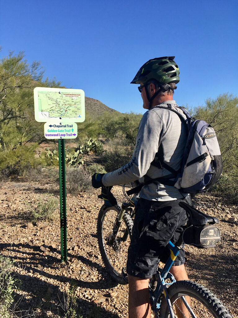 Man in bike helmet, long sleeved grey shirt and grey camouflage biking shorts standing over mountain bike looking at trail sign.