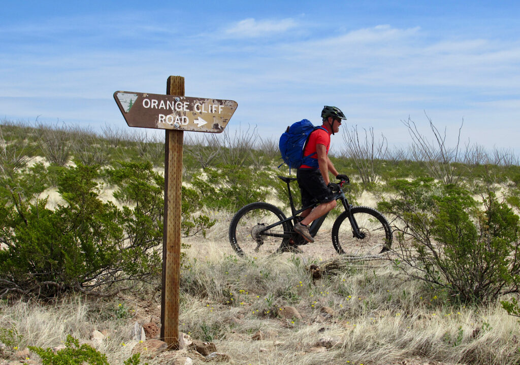Man in red shirt and black shorts with blue backpack riding mountain bike behind sign reading: Orange Cliff Road.