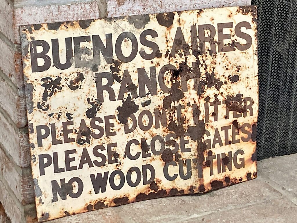 Rusting metal sign painted white with words: BUENOS AIRES RANCH PLEASE DON'T LITTER PLEASE CLOSE GATES NO WOOD CUTTING.