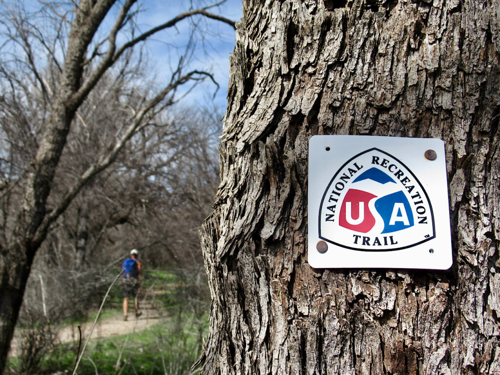 Man in background with blue backpack hiking trail through the woods. Sign on tree trunk in foreground reads: National Recreation Trail with letters USA in red, white and blue.