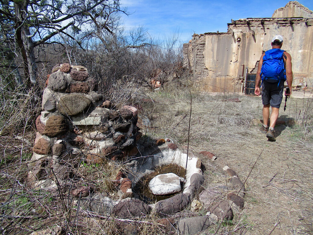 Man in shorts wearing blue backpack walking through abandoned ranch house yard with old rock fountain in foreground.
