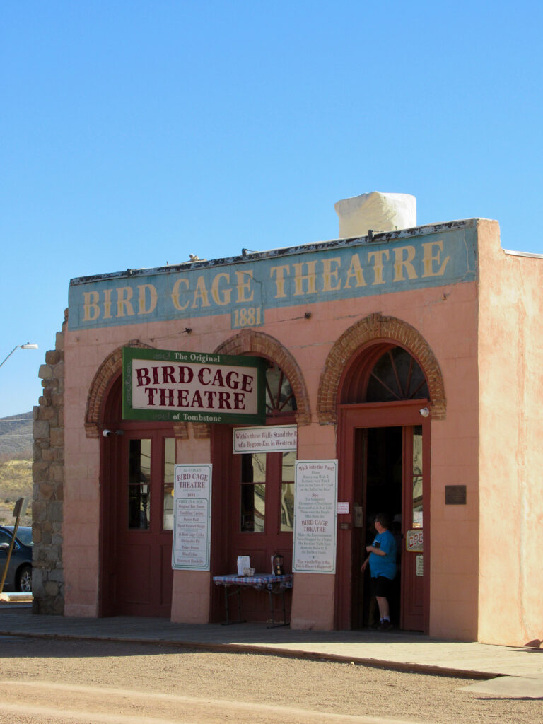 Pink adobe building with three arched doorways. Man standing in righthand doorway. Sign above painted in blue reads: Bird Cage Theatre.