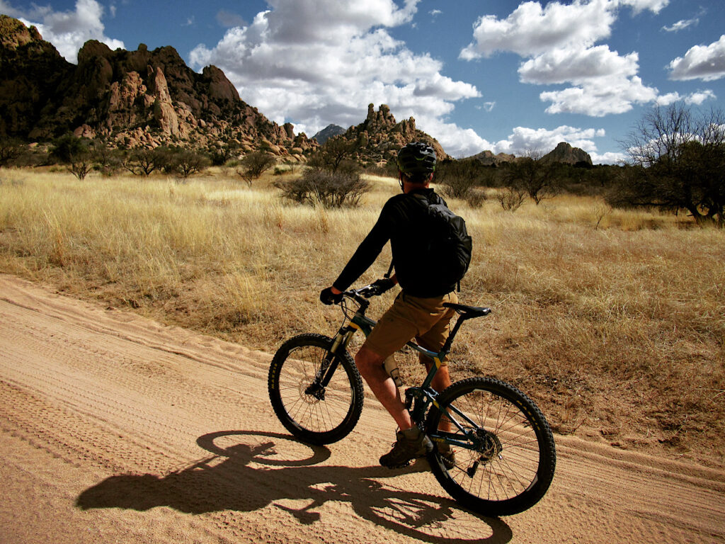 Man standing astride a mountain bike on a dirt road looking out over grassy field towards brown sandstone boulders and mountains.