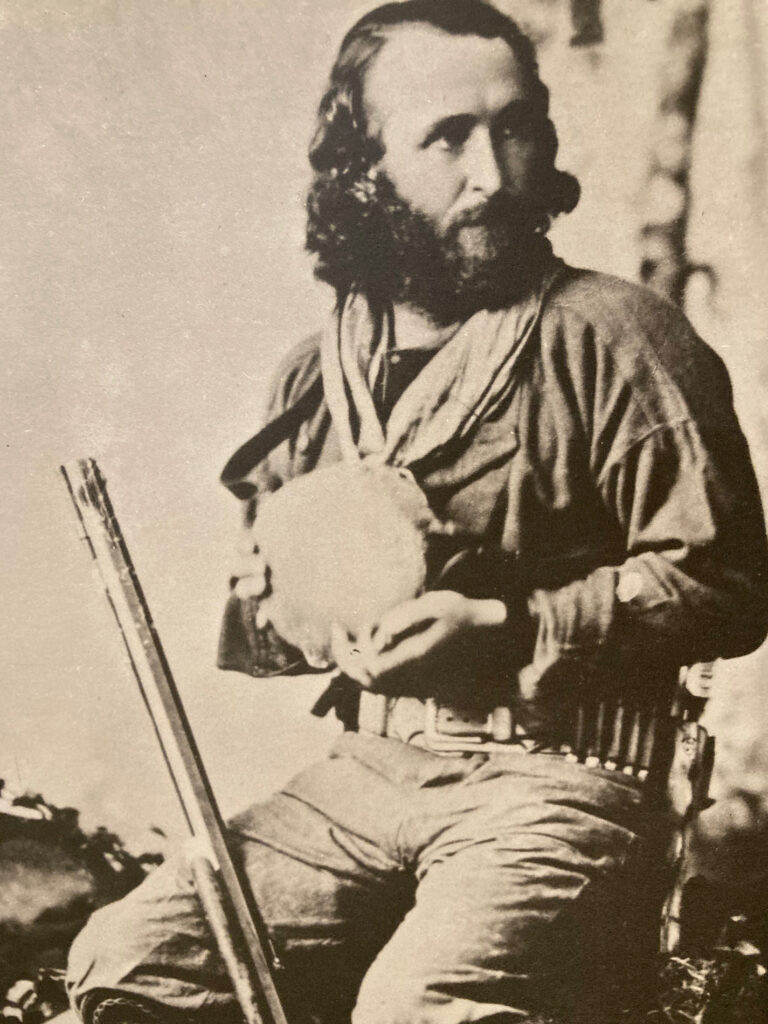 Black and white photo of a man with long hair and beard holding a canteen with a rifle propped against his leg.