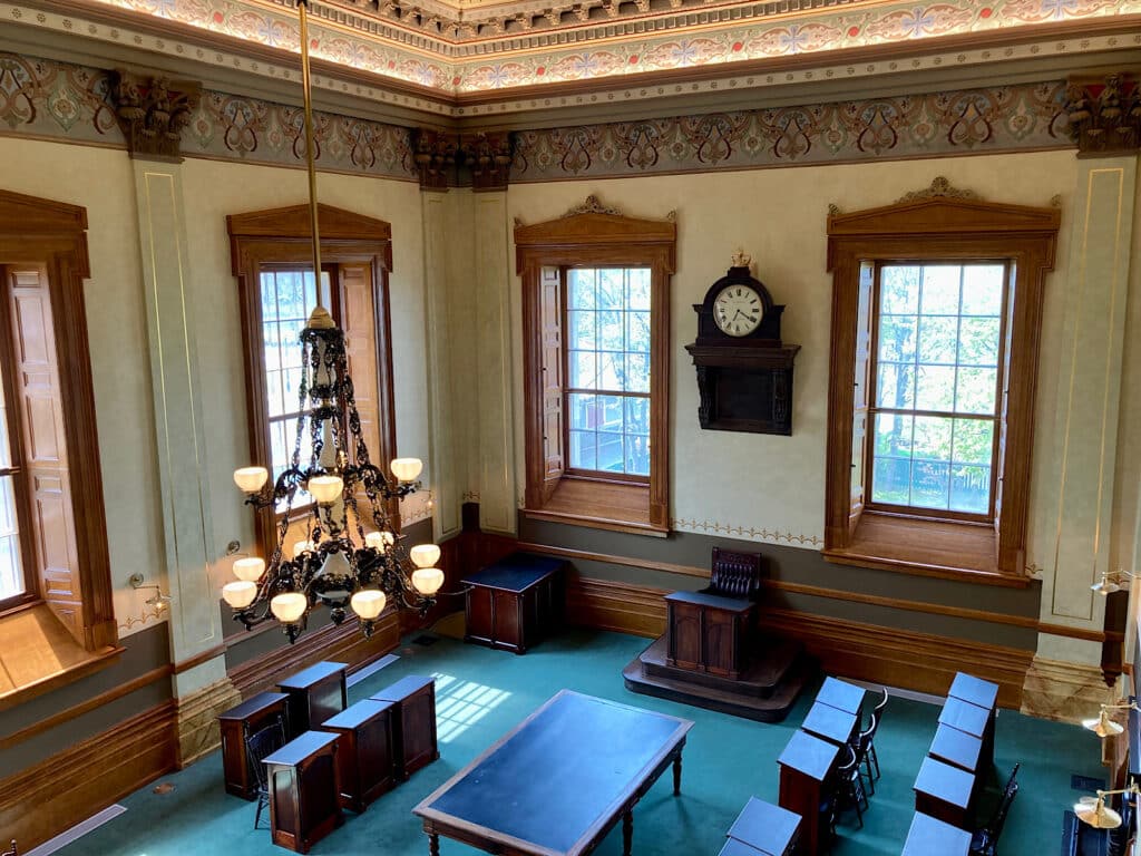 Large legislative room with wooden desks in rows, overweight windows, a large chandelier and wall clock.
