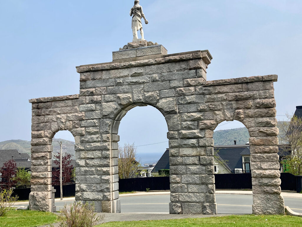 Stone triple arch with statue in middle on top looking out to view of buildings and a distant harbour.