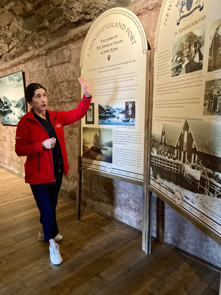 Woman in red coat, black pants and top standing by interpretive signage with left arm raised.