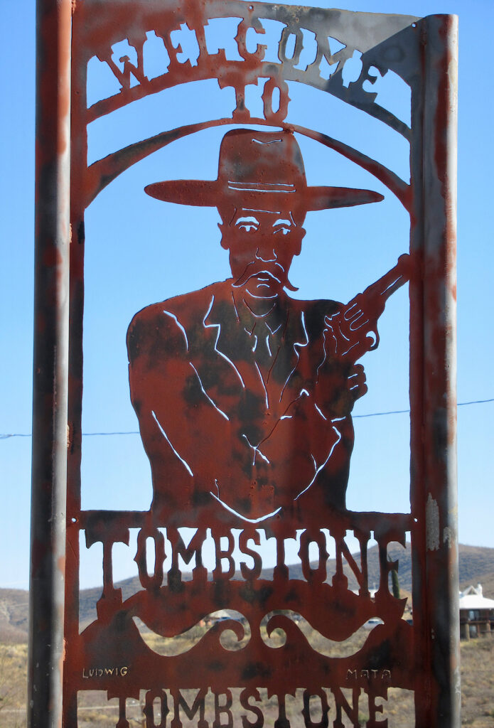 Metal cutout sign with image of a moustached man wearing a flat brim hat and hold up a gun in the middle of the words "Welcome to Tombstone"