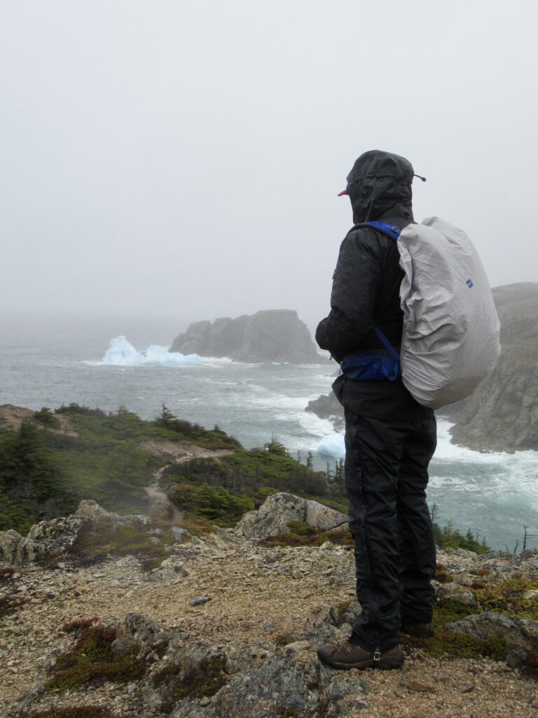Man in black rain gear with grey pack cover standing on cliff overlooking ocean and icebergs.