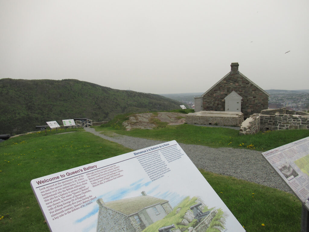 Interpretive sign with title "Welcome to Queen's Battery overlooking gravel path, lawn and a stone building and foundations.