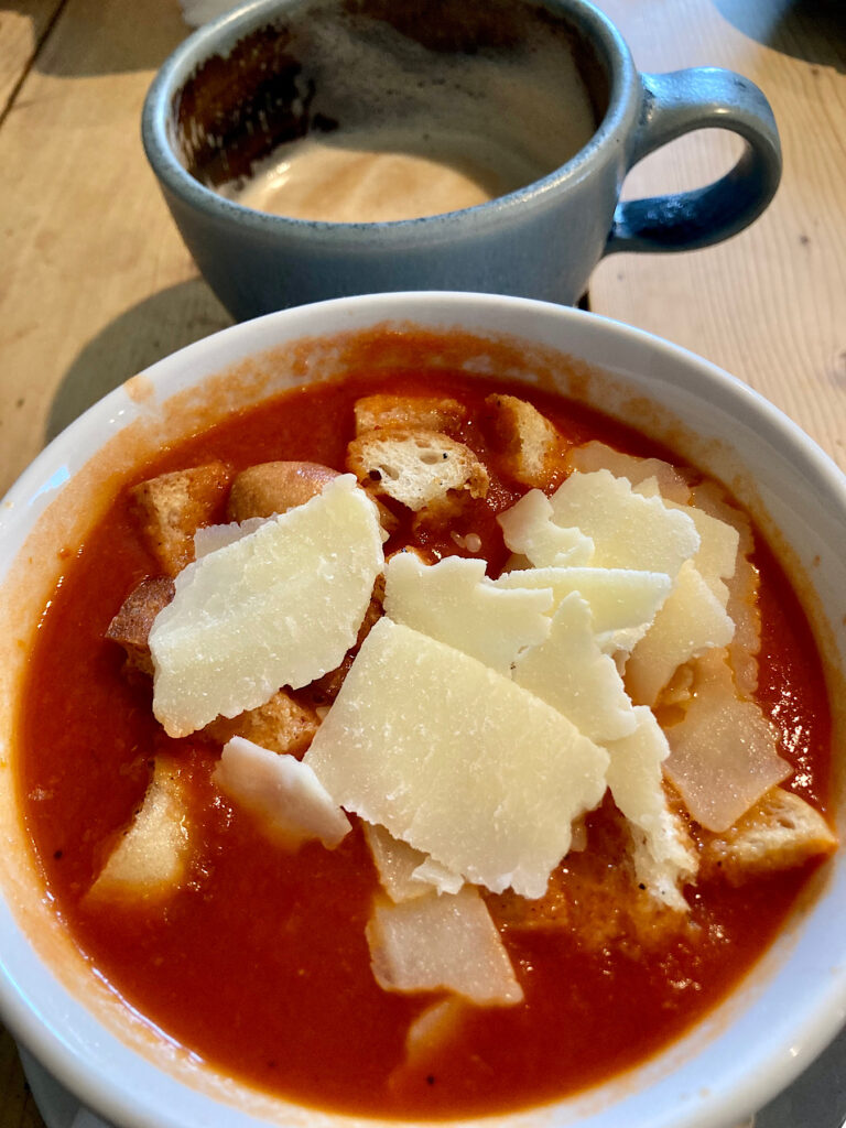 Bowl of tomato soup with chunks of white cheese on top and large latter in pottery mug.