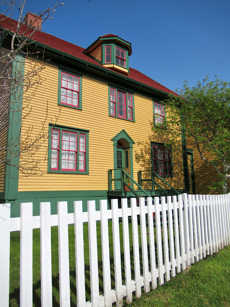 White picket fence in front of two-storey home painted gold with dark green trim.
