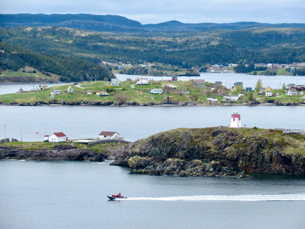 Boat in ocean in front of lighthouse and distance houses in a bay.