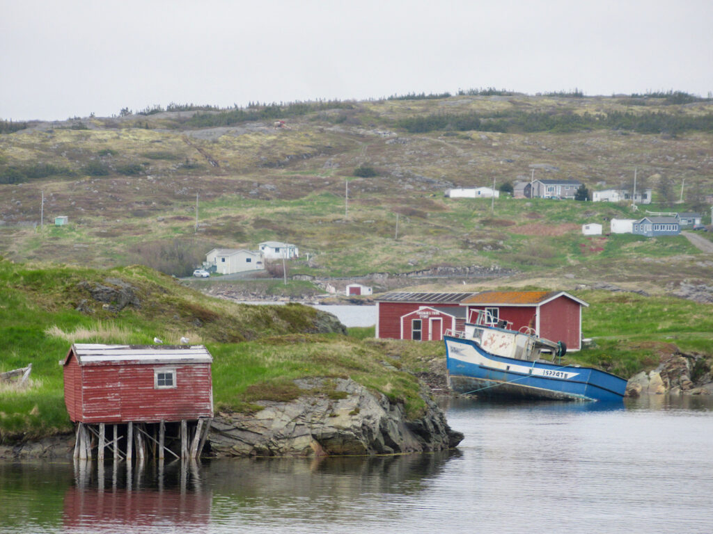 Red painted wooden buildings on stilts at water's edge with blue and white fishing boat tucked in rocky bay.