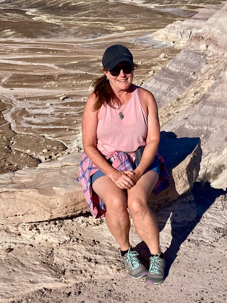 Woman in black hat and sunglasses, pink shirt and blue shorts sitting on rock at edge of buff-coloured mesa.