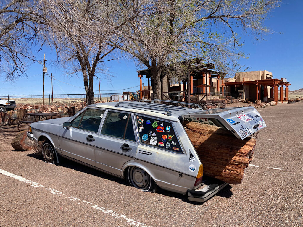 Old silver wagon with two flat tires and large piece of petrified wood hanging out of the back in front of gift shop.