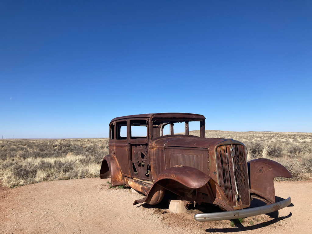 Historic rusting body of a old car sitting on the edge of a field of dried grasses and low shrubs under blue sky.