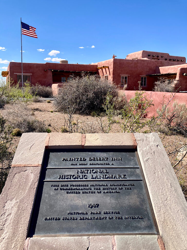 Bronze plaque reading Painted Desert Inn National Historic Landmark in front of a low pink adobe building with American flag flying under blue sky.