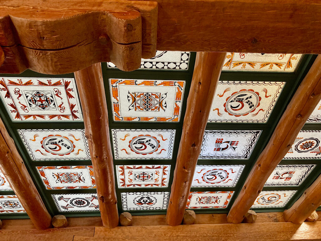 Wooden ceiling with large beams separated by painted glass squares with black, red, orange and brown Native American designs.