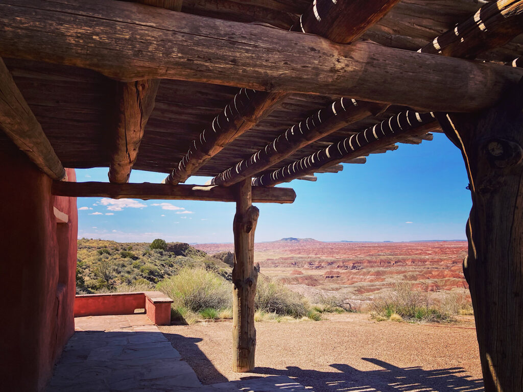 Wooden covered porch overlooking distant red hills below under blue sky.