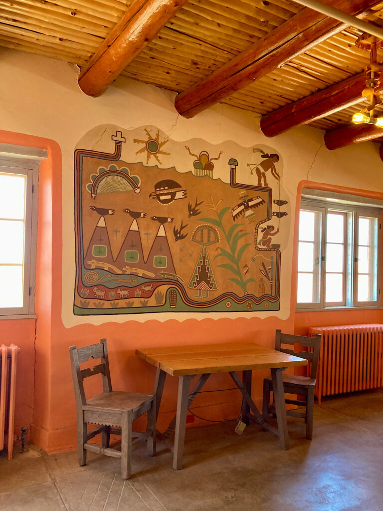 Painted mural on wall sits between two windows, below a wooden ceiling and above a table with two grey chairs that sit on a concrete floor.