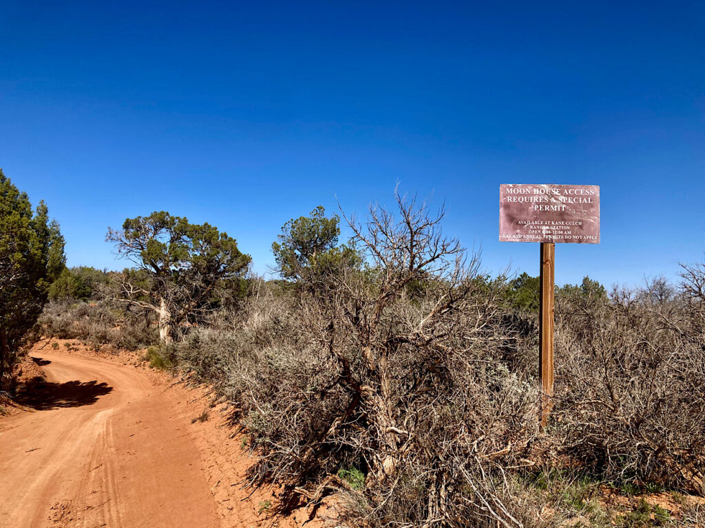 Brown sign with white lettering reading "Moon House access requires special permit" standing to right of dirt track in middle of scrub forest under blue sky.