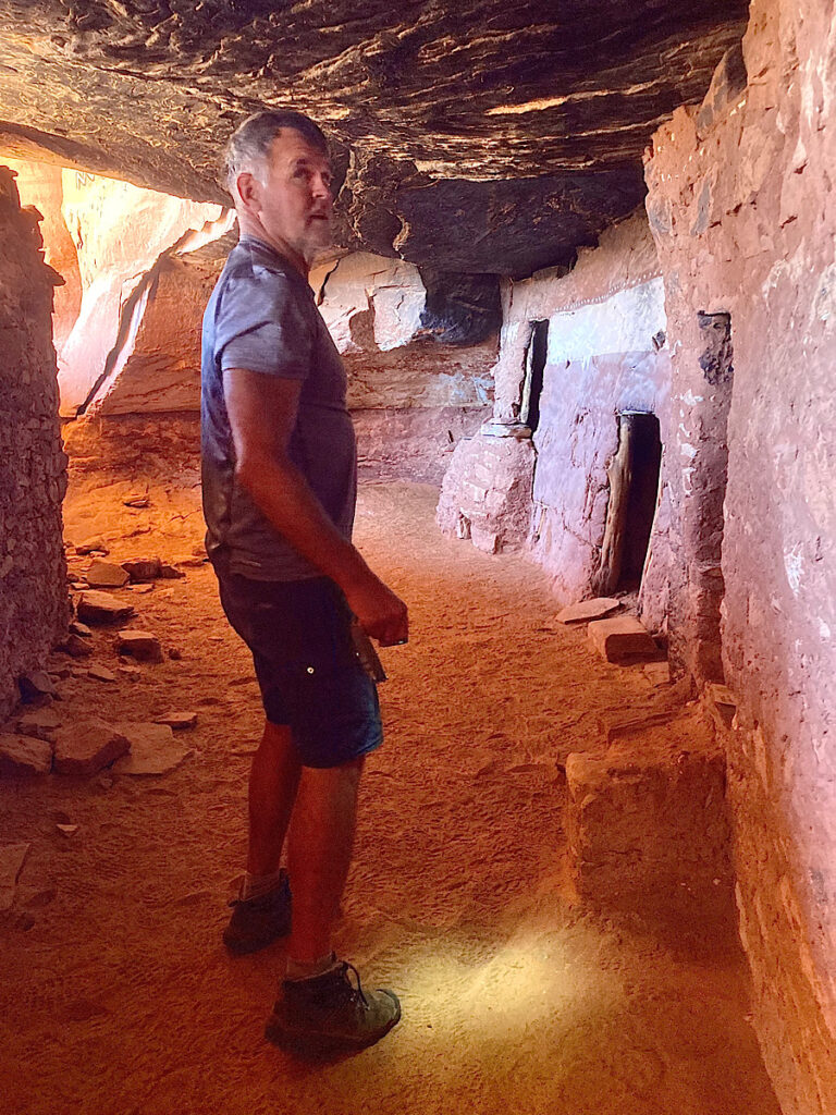Man in grey shorts and t-shirt wearing hiking boots inside a cave with stone-built rooms.