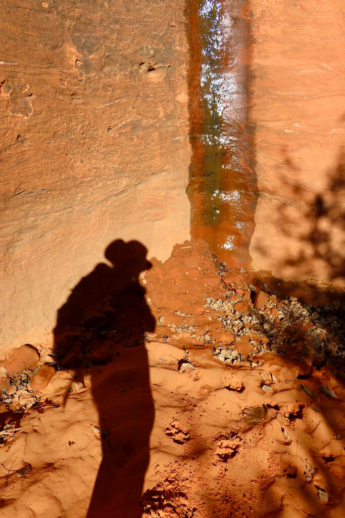 Shadow of a woman with backpack to one side of a thin waterfall on smooth red sandstone.
