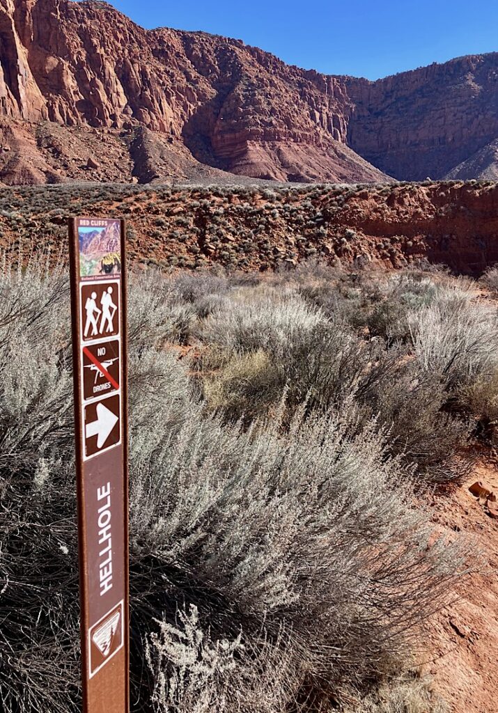 Trail sign marking Hellhole Canyon with tall red cliffs in background.