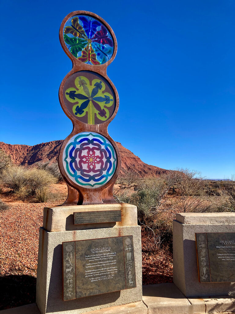 Glass and metal sculpture with red rocks in background under blue sky.