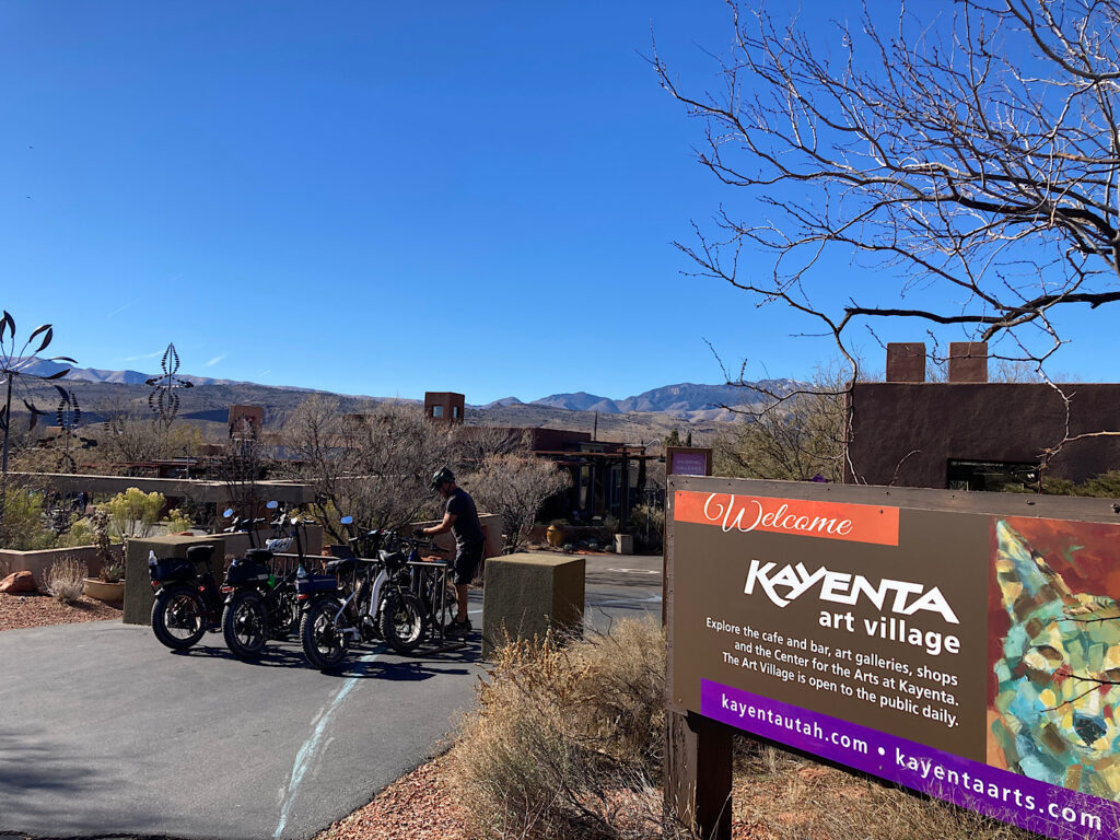 Sign reading: Welcome Kayenta Art Village with man in background locking up a bike in bike stand.