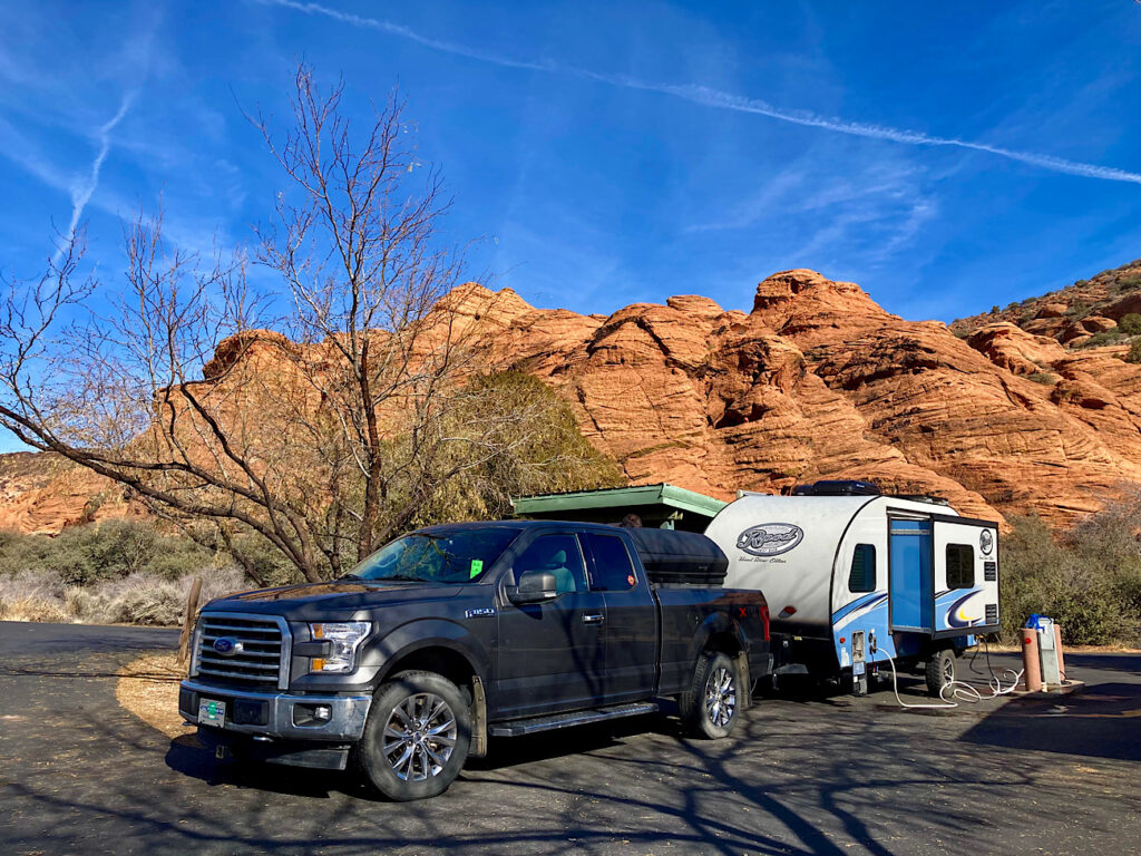 Grey pickup truck and blue and white trailer set up in campsite with red rock cliffs in background beneath blue sky.