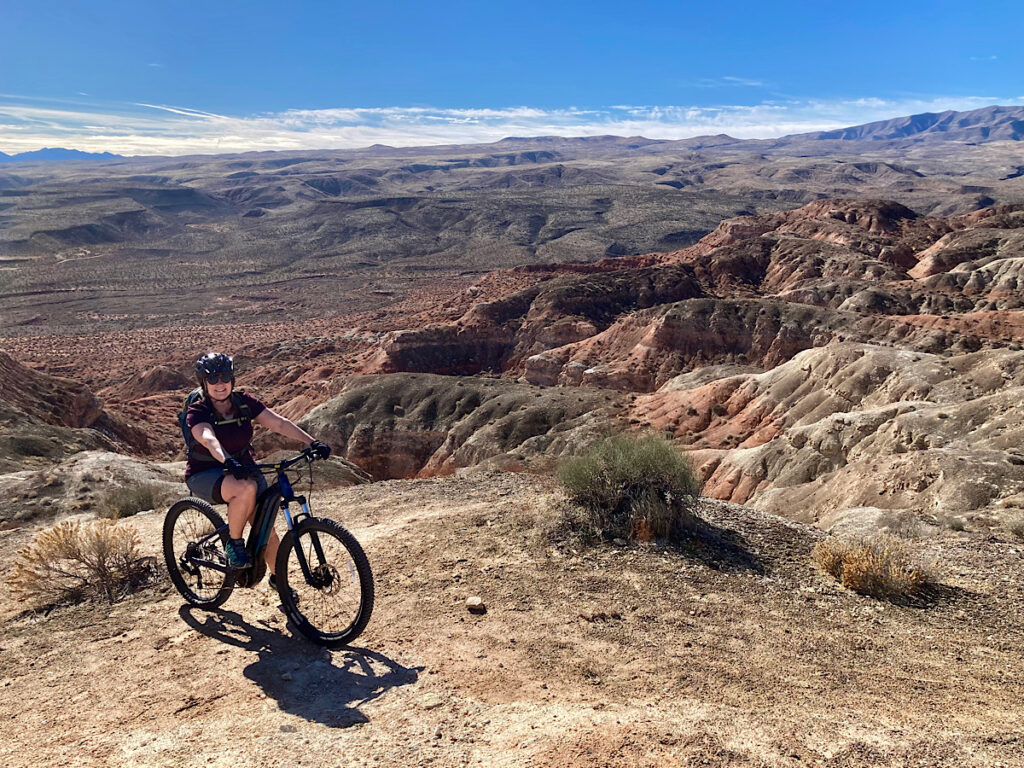 Woman in shorts on a mountain bike with dramatic red rock canyon country in background under blue sky.