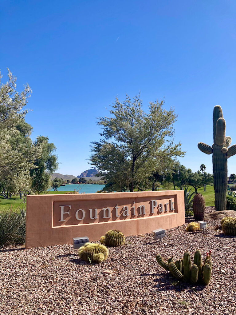 Long brown concrete sign reading "Fountain Park" in front of cactus garden with lake in background under blue sky.