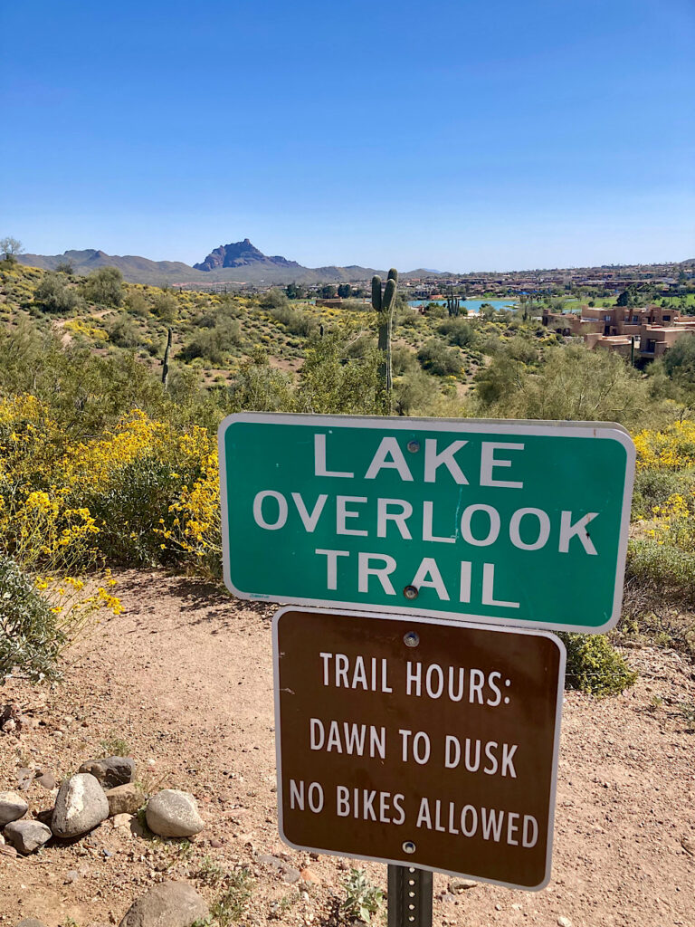 Desert hillside with distant lake and small peak behind a green sign with white lettering reading: Lake Overlook Trail. Brown sign with white lettering below reading: Trail Hours: Dawn to Dusk No Bikes Allowed