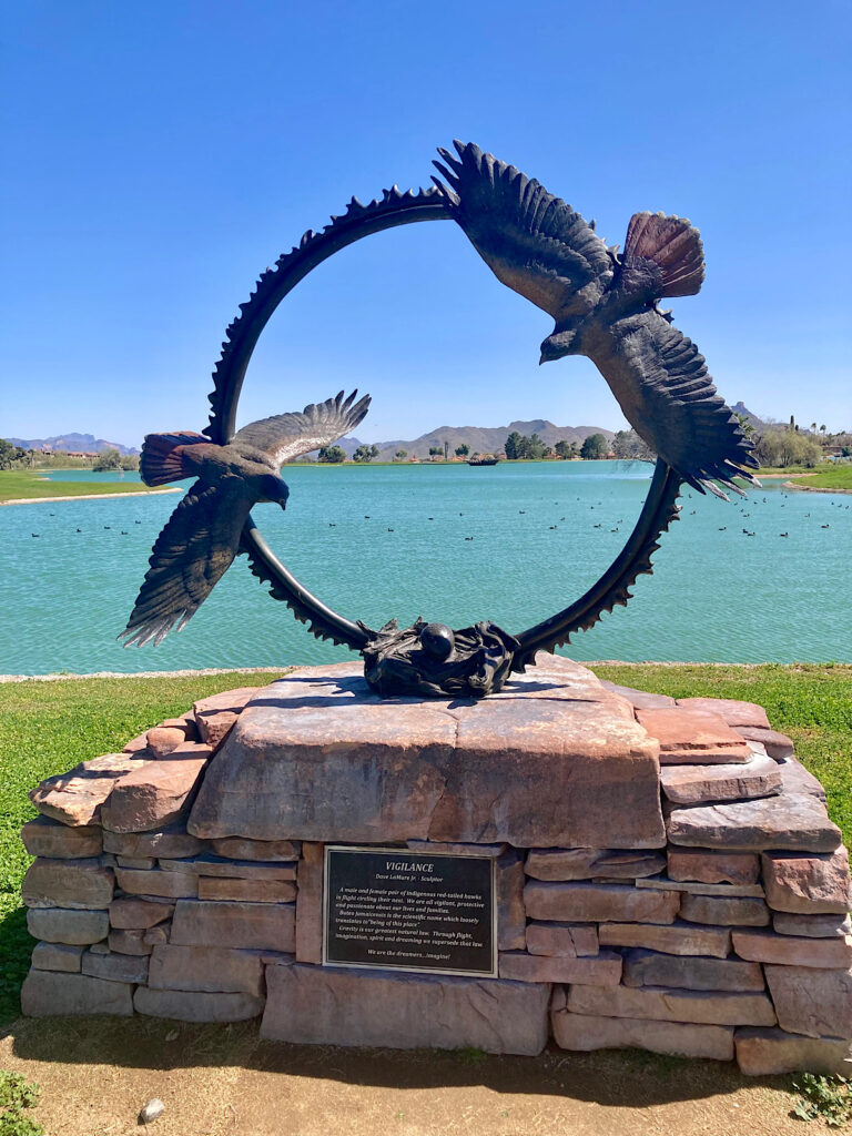 Circular bronze sculpture with two hawks above a next on a stone pedestal set in front a blue-green lake.