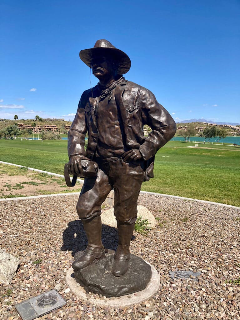 Bronze sculpture of moustached man stand in knee high boots, pants, shirt, jacket and hat holding a water canteen in right hand.