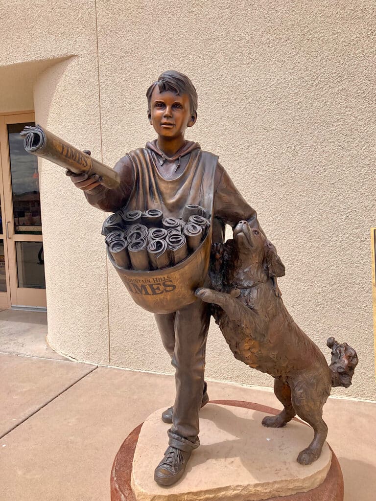 Bronze sculpture of young newspaper delivery boy holding paper out while petting dog who has jumped up on him.
