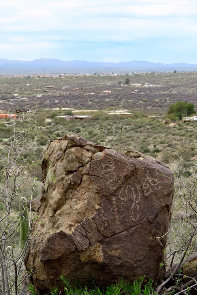 Large boulder in foreground inscribed with waving lines. Homes in distance below slope. 