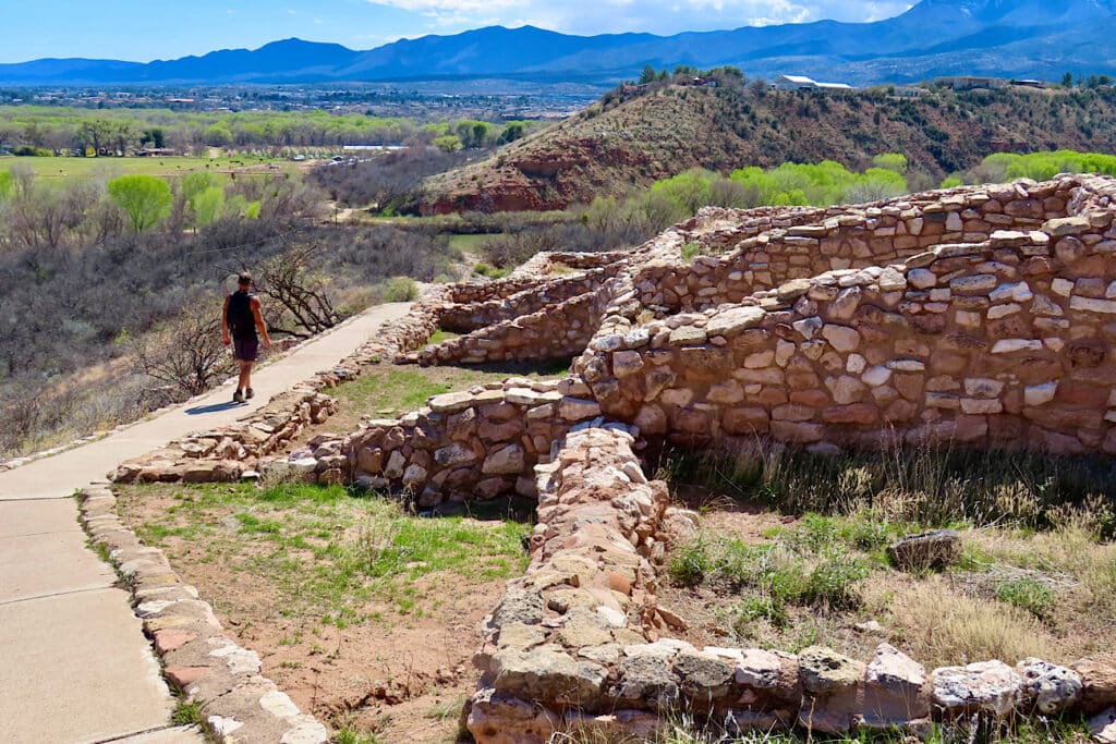 Man in dark shorts and short sleeved top walking paved path by stone walls on hill above green valley with dark mountains in background.