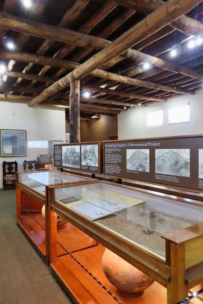 Interior of a museum with large dark wood beams on ceiling, white walls and glass display cases with clay pots and interpretive signage.