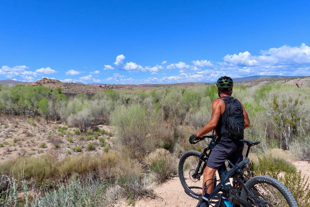 Man wearing dark short and short sleeved top standing astride a mountain bike overlooking a valley and low hills.