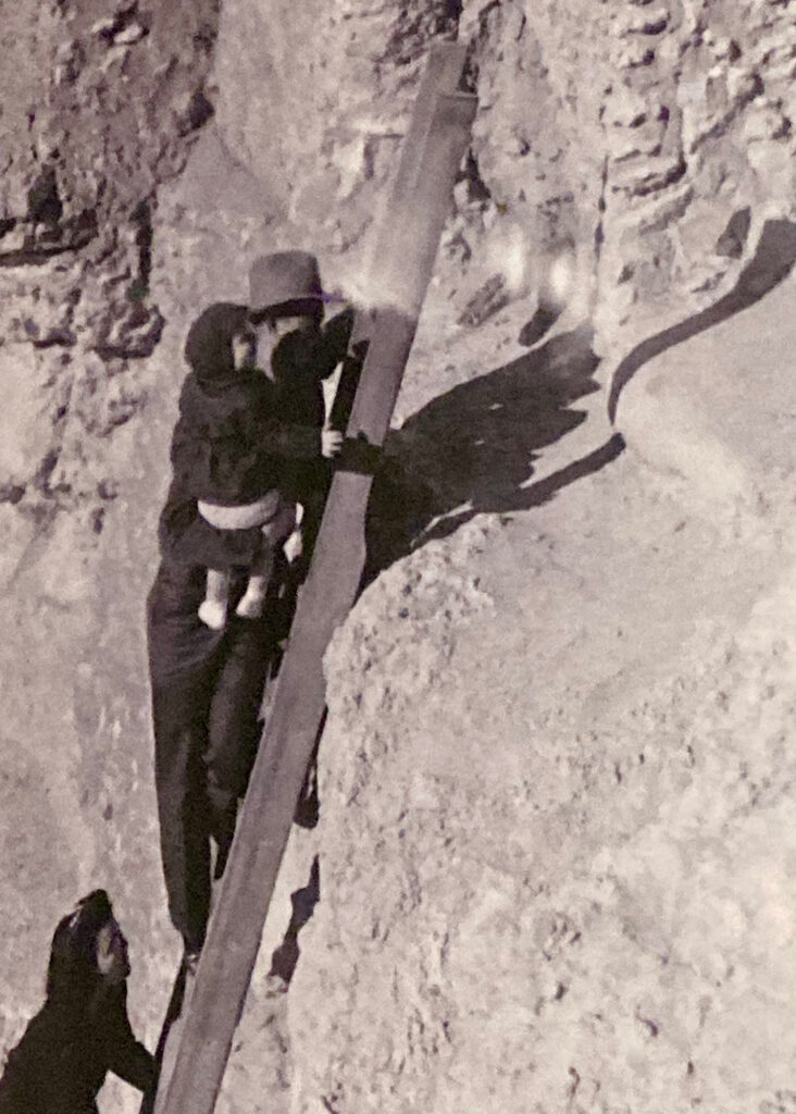 Black and white photo of man holding a child while climbing a ladder on a cliff with a woman coming up below.