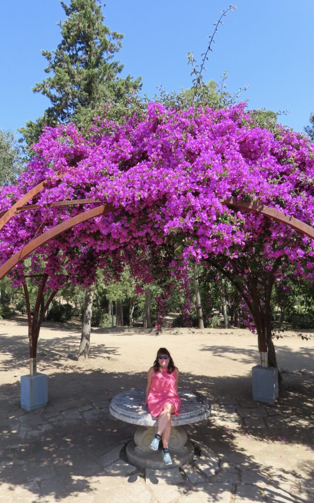 Purple flower trellis under blue sky with woman in pink dress sitting on round stone table below.