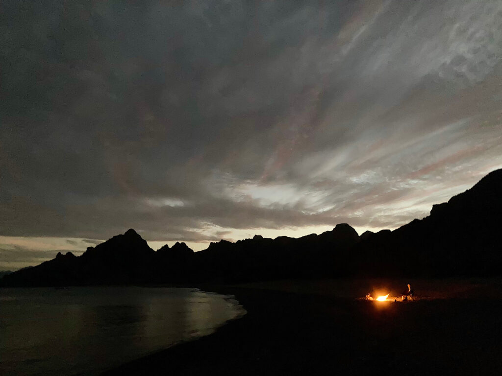 Sunset silhouette of mountains with orange glow of distant campfire.