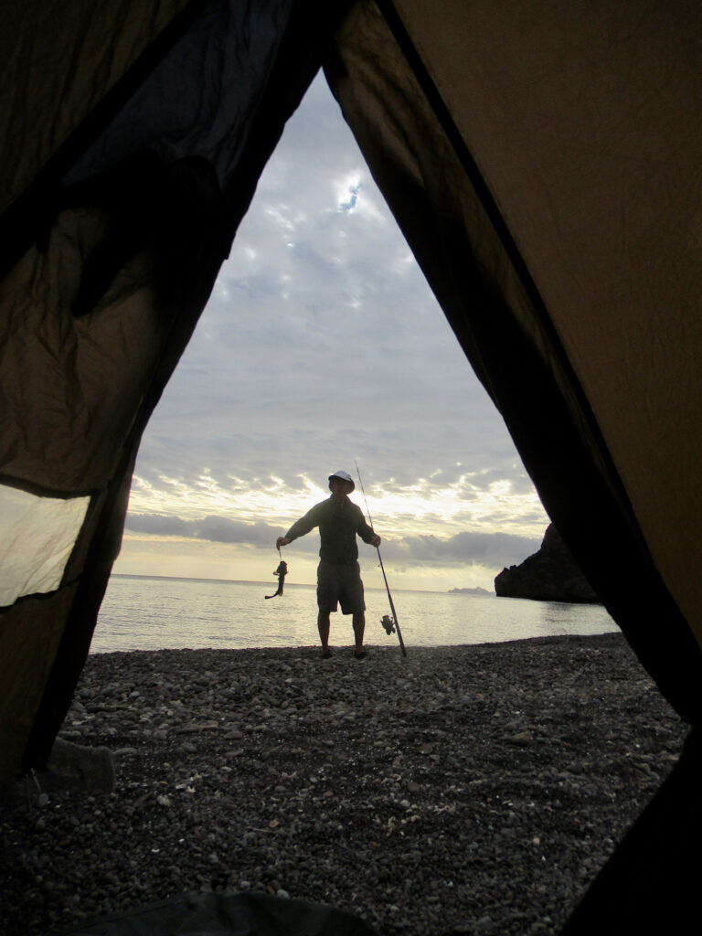 Man silhouetted in front of tent opening while holding a fish on a string.