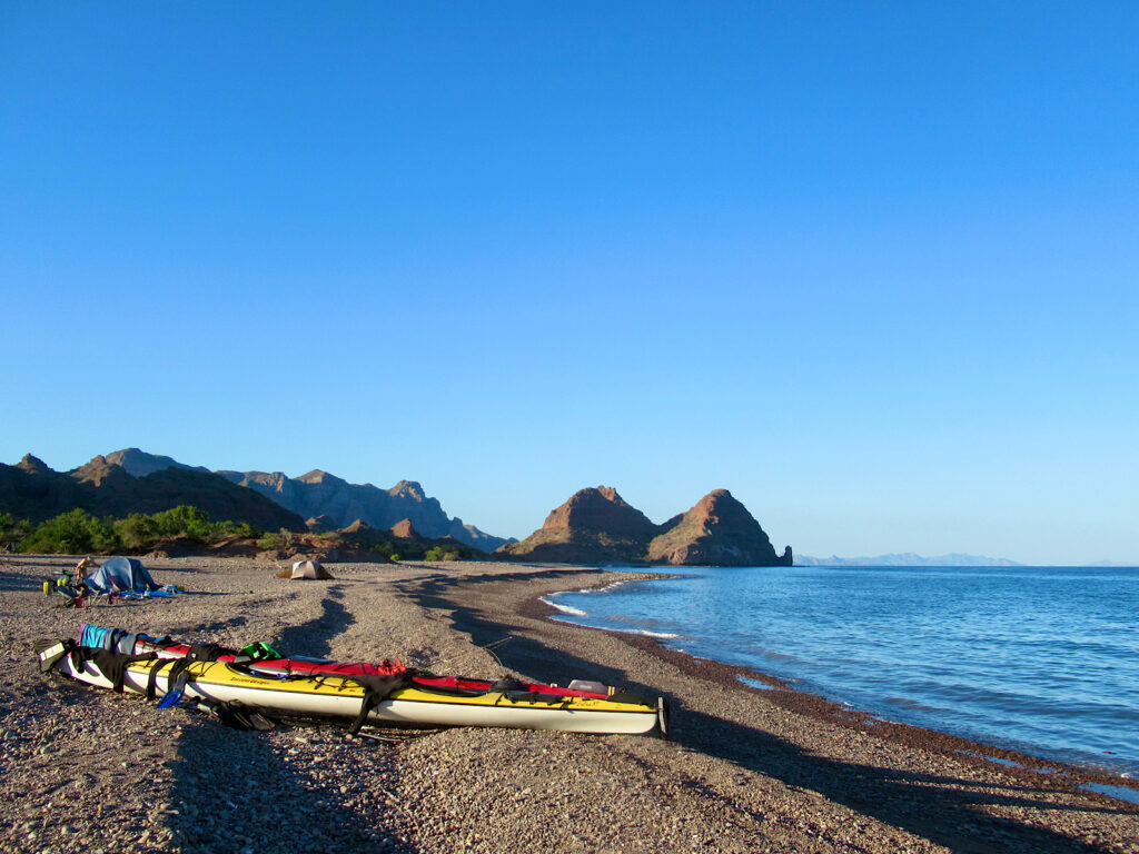 Yellow and red tandem kayaks pulled up on beach in front of two tents with distant mountains under blue sky.