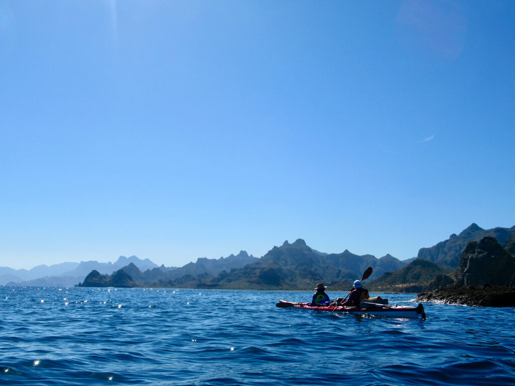 Two people in red tandem kayak on water in front of rugged, mountainous coastline.