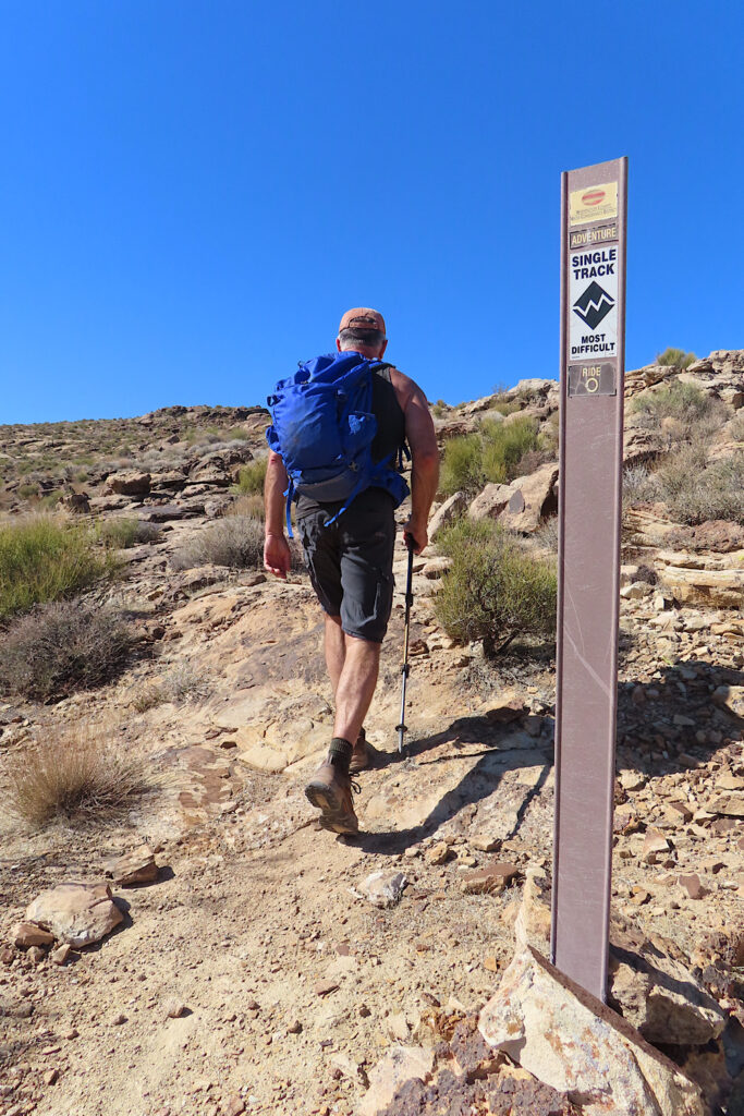 Man in shorts and backpack hiking up a biking trail signed: Adventure trail